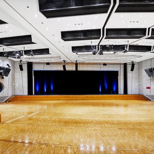 Innenansicht auf den unbestuhlten Kombisaal. Die Wände sind in hellen Tönen gehalten, der Saal verfügt über einen Parkettboden. Hinten im Saal ist eine Bühne zu sehen.