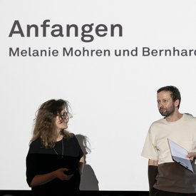 Kuratorin Melanie Mohren und Kurator Bernhard Herbordt während ihrer Eröffnungsrede. Im Hintergund auf der Leinwand: »Anfangen. Melanie Mohren und Bernhard Herbordt«.