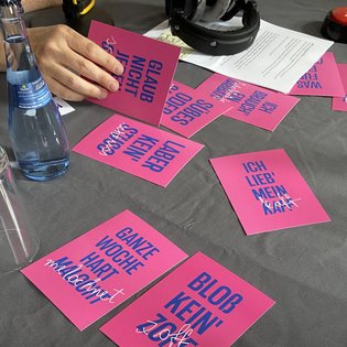 Auf einem Tisch mit einer grauen Tischdecke stehen verschiedene Getränke und es liegen pinke Postkarten aus dem Projekt »Schlamassel Tov« aus.