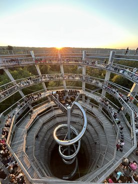 Menschen sitzen und stehen auf spiralförmigen Holzstegen eines Baumwipfelpfads mit zentralem Metallrutschenturm bei Sonnenuntergang