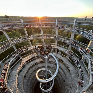 Menschen sitzen und stehen auf spiralförmigen Holzstegen eines Baumwipfelpfads mit zentralem Metallrutschenturm bei Sonnenuntergang