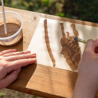 Person malt mit einem Pinsel auf einem Blatt Papier, das auf einem Holztisch liegt. Der Pinsel wird in braune Farbe getaucht, die sich in einem Plastikbecher befindet. Im Hintergrund ist verschwommenes Grün zu erkennen, was auf eine kreative Tätigkeit im Freien hinweist.