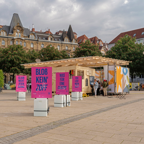 Foto der Ausstellung »Schlamassel Tov« auf dem Marienplatz vor der Sukka: Mehrere pinke Plakate mit blauer Aufschrift stehen vor einem Bau aus Holz und Stoff – der Laubhütte bzw. Sukka von Sukkat Salaam. Im Vordergrund ist ein Plakat mit der Aufschrift »Bloß kein‘ Zoff« zu sehen.