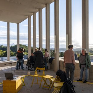 Terrasse des Literaturmuseum der Moderne auf der Schillerhöhe. Im Vordergrund stehen gelbe Stühle und Tischchen im Hintergrund sind mehrere Personen miteinander im Gespräch. Die Abendsonne scheint durch die Säulen der Terrasse hindurch.