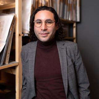 Mann mit Brille, dunklem Rollkragenpullover und grauem Sakko lehnt an einem Regal mit großen, verpackten Büchern oder Kunstwerken in einem Raum mit grauer Wand