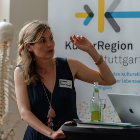 Nicola Bramkamp sitzt in einem dunklen, ärmellosen Kleid vor einem Banner mit der Aufschrift ‚KulturRegion Stuttgart‘. Mit erhobener Hand gestikuliert sie, während die andere Hand auf einem Tisch mit Flasche und Papieren ruht. Im Hintergrund ist ein Skelettmodell zu sehen.