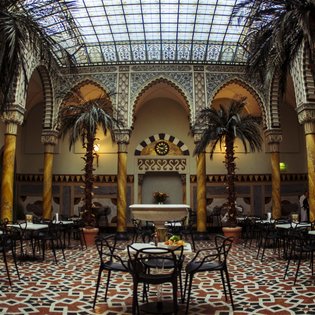 Innenraum der Maurischen Halle im Palais Thermal Bad Wildbad mit Säulen, Palmen in Töpfen und Tischen mit Stühlen auf einem gemusterten Boden unter einem Glasdach