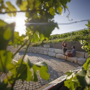 Zwei Personen, möglicherweise ein Paar sitzen auf halbrund angeordneten Steinen. Es wurde durch eine Weinrebe hindurch fotografiert, so dass im Vordergrund am Bildrand noch verschwommen Blätter erkennbar sind