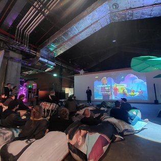 Teilnehmer des Symposiums in Stuttgart betrachten eine bunte Leinwandpräsentation zu Kunst im Gaming-Kontext.