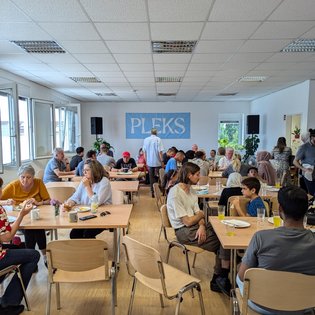 Begegnungscafe in den Räumlichkeiten des PLEKS. An verschiedenen Tischen sitzen Menschen zusammen, essen, trinken und unterhalten sich.