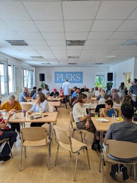 Begegnungscafe in den Räumlichkeiten des PLEKS. An verschiedenen Tischen sitzen Menschen zusammen, essen, trinken und unterhalten sich.