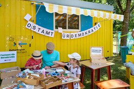 vor einem gelben Bauwagen mit der Aufschrift "Postamt für Demokratie" sitzen Menschen auf Holzbänken, Erwachsene und Kinder und basteln am Tisch