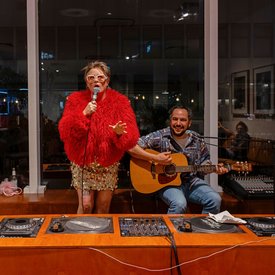 Person mit rotem, flauschigem Pullover und goldfarbenem Rock singt mit Mikrofon neben sitzendem Gitarristen in gemustertem Hemd vor DJ-Pult in Innenraum mit großen Fenstern