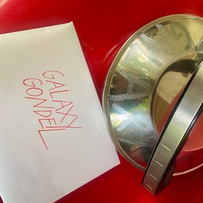 Ältere, metallische Teekanne mit Griff auf rotem Untergrund neben einem weißen Zettel mit handschriftlicher roter Aufschrift 'GALAXY GONDEL'