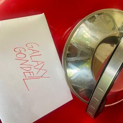 Ältere, metallische Teekanne mit Griff auf rotem Untergrund neben einem weißen Zettel mit handschriftlicher roter Aufschrift 'GALAXY GONDEL'