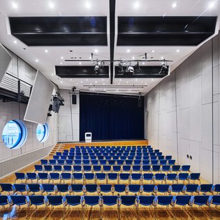 Innenansicht auf den bestuhlten kleinen Saal. Die Wände sind in hellen Tönen gehalten, der Saal verfügt über einen Parkettboden. Hinten im Saal ist eine Bühne zu sehen