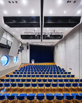 Innenansicht auf den bestuhlten kleinen Saal. Die Wände sind in hellen Tönen gehalten, der Saal verfügt über einen Parkettboden. Hinten im Saal ist eine Bühne zu sehen