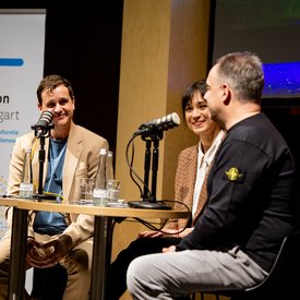 Moderator Ingmar Volkmann, Rocío Alemán und Alexander Zorniger während der Podcast Aufnahme. Alle drei sitzen auf Barhockern an Tischen und haben Mikrofone vor sich. 