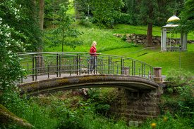 Frau in rotem Oberteil steht auf einer geschwungenen Steinbrücke mit Metallgeländer in einem grünen Park mit Bäumen und Pavillon