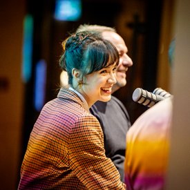 Rocío Alemán und Alexander Zorniger während der Podcast Aufnahme. Sie sitzen beide auf Barhockern und haben Mikrofone vor sich. Der Fokus der Aufnahme liegt auf Rocío Alemán, Alexander Zorniger ist etwas verdeckt zu sehen.