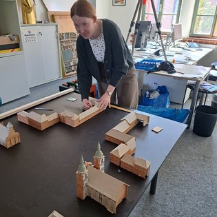 Frau mit Zopf und Strickjacke baut auf einem Tisch ein Architekturmodell aus Holz und Pappe in einem hellen Raum mit Schreibtischen und Fenstern
