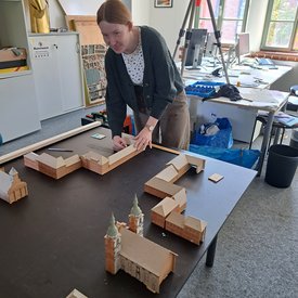 Frau mit Zopf und Strickjacke baut auf einem Tisch ein Architekturmodell aus Holz und Pappe in einem hellen Raum mit Schreibtischen und Fenstern