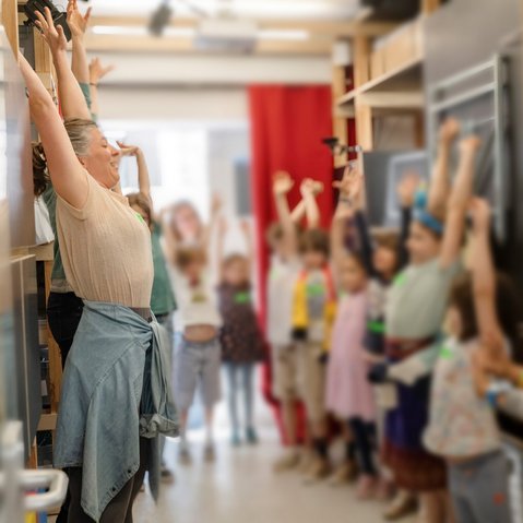 Eine Gruppe Kinder strecken ihre Arme offensichtlich nach Anleitung einer erwachsenen Person am linken Bildrand Richtung Decke.