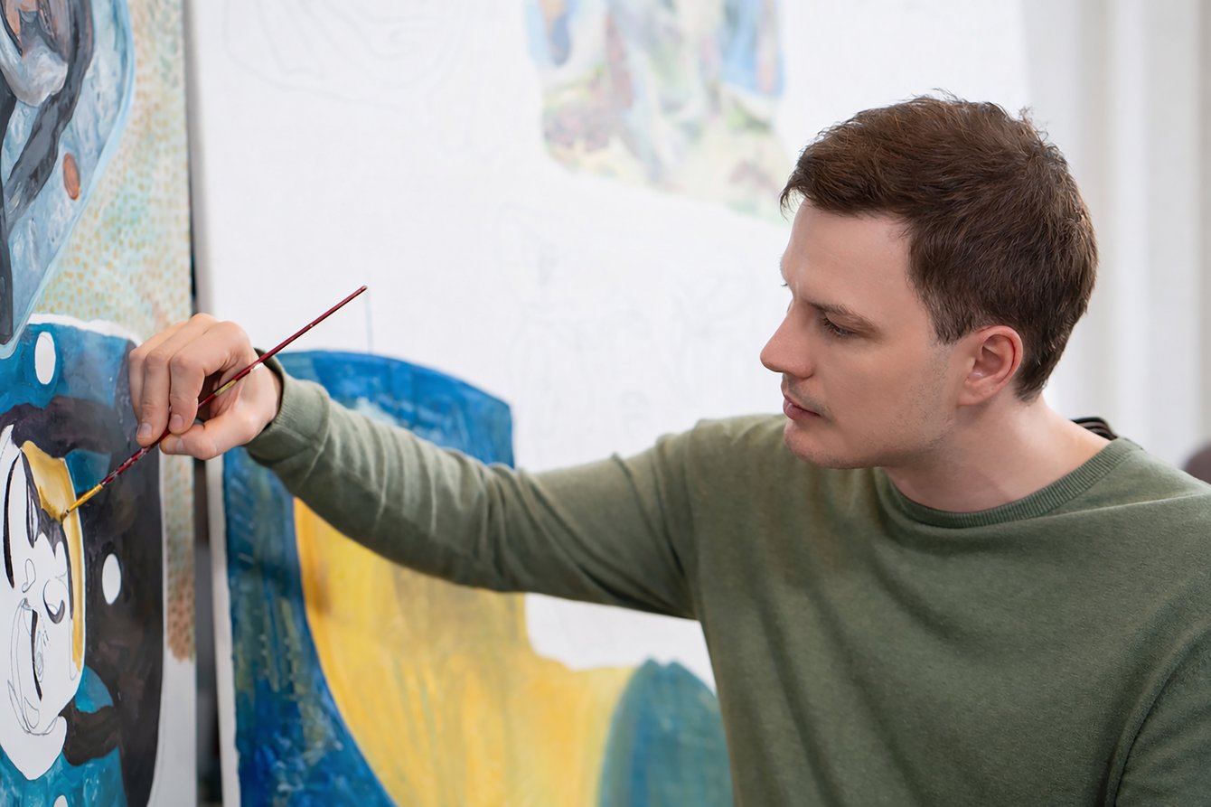 Person mit grünem Pullover malt mit Pinsel an einer farbigen Leinwand.