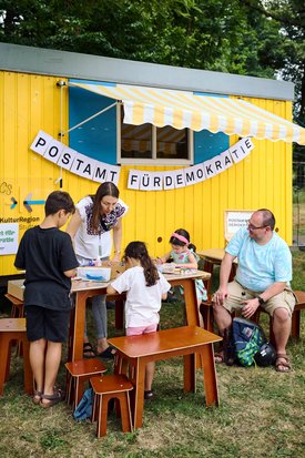 vor einem gelben Bauwagen mit der Aufschrift "Postamt für Demokratie" sitzen Menschen auf Holzbänken, Erwachsene und Kinder und basteln am Tisch
