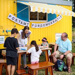 vor einem gelben Bauwagen mit der Aufschrift "Postamt für Demokratie" sitzen Menschen auf Holzbänken, Erwachsene und Kinder und basteln am Tisch