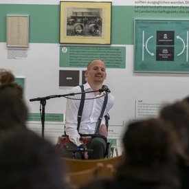 Blick auf Michael Turinsky beim Salon in Böblingen – über die Köpfe der Zuschauer*innen hinweg. Turinsky trägt eine dunkle Hose und ein weißes Hemd mit Hosenträgern. Er sitzt vor einem Mikrofon und nutzt einen Rollstuhl.