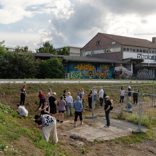 Menschen arbeiten gemeinsam in einem städtischen Freiraum mit Gr Gruppe an Menschen auf einem leicht abschüssigen, grasbewachsenen Gelände – teils knieend. Die Gruppe sammelt Erde, die später im Workshop zu Farbe verarbeitet wird.. Ein Schild mit der Aufschrift ‚Zollamt‘ ist am Gebäude zu sehen