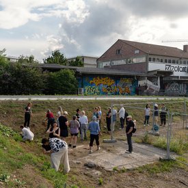 Menschen arbeiten gemeinsam in einem städtischen Freiraum mit Gr Gruppe an Menschen auf einem leicht abschüssigen, grasbewachsenen Gelände – teils knieend. Die Gruppe sammelt Erde, die später im Workshop zu Farbe verarbeitet wird.. Ein Schild mit der Aufschrift ‚Zollamt‘ ist am Gebäude zu sehen