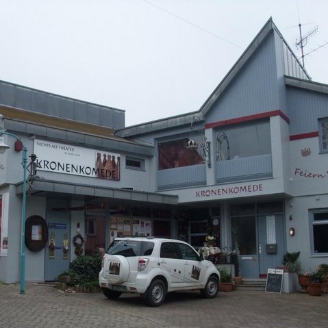 Außenansicht des Gebäudes „KronenKomede“ in Filderstadt, Deutschland. Es handelt sich um ein Theaterhaus mit der Aufschrift „NICHTS ALS THEATER“ und „Feiern Sie Ihre Feste hier“. Ein weißes Auto mit dem Logo des Theaters steht vor dem Eingang. Die Fassade ist hellgrau mit roten Akzenten, dekoriert mit Theatermotiven und Pflanzen.