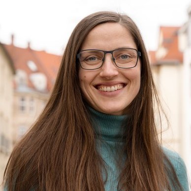 Portrait von Tina Pantisano. Sie hat langes, glattes, braunes Haar, trägt eine Brille und einen türkisfarbenen Rollkragenpulli. Tina Pantisano lächelt freundlich in die Kamera.