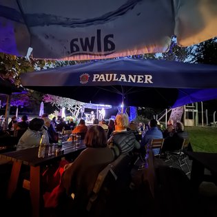 Drei Musiker treten abends auf einer kleinen Außenbühne auf. Links spielt ein Mann Akkordeon, in der Mitte und rechts singen zwei Männer ins Mikrofon. Die Bühne ist von Dunkelheit umgeben, im Vordergrund sitzen Zuschauer im Schatten.