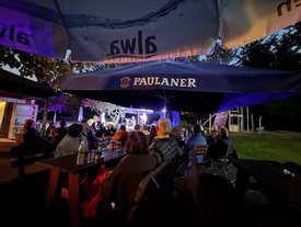 Drei Musiker treten abends auf einer kleinen Außenbühne auf. Links spielt ein Mann Akkordeon, in der Mitte und rechts singen zwei Männer ins Mikrofon. Die Bühne ist von Dunkelheit umgeben, im Vordergrund sitzen Zuschauer im Schatten.