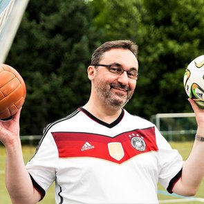 Mann mit Brille und schwarz-weißem Shirt steht vor einem Fußballtor im Freien und hält zwei Fußbälle in den Händen. Er lächelt und blickt direkt in die Kamera.