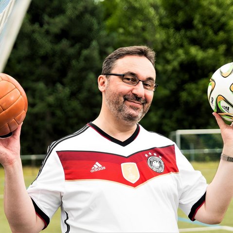 Mann mit Brille und schwarz-weißem Shirt steht vor einem Fußballtor im Freien und hält zwei Fußbälle in den Händen. Er lächelt und blickt direkt in die Kamera.