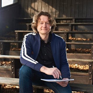 Person mit Locken sitzt auf Holztribüne im Freien, trägt blauen Trainingsanzug, schreibt auf Papier. Herbstliche Umgebung mit Laub.