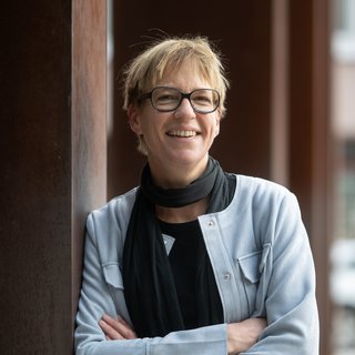 Eva Wolfangel: Person lehnt mit einer Hand in der Hosentasche an einer Wand und trägt eine helle Jacke, schwarzen Schal und schwarze Hose