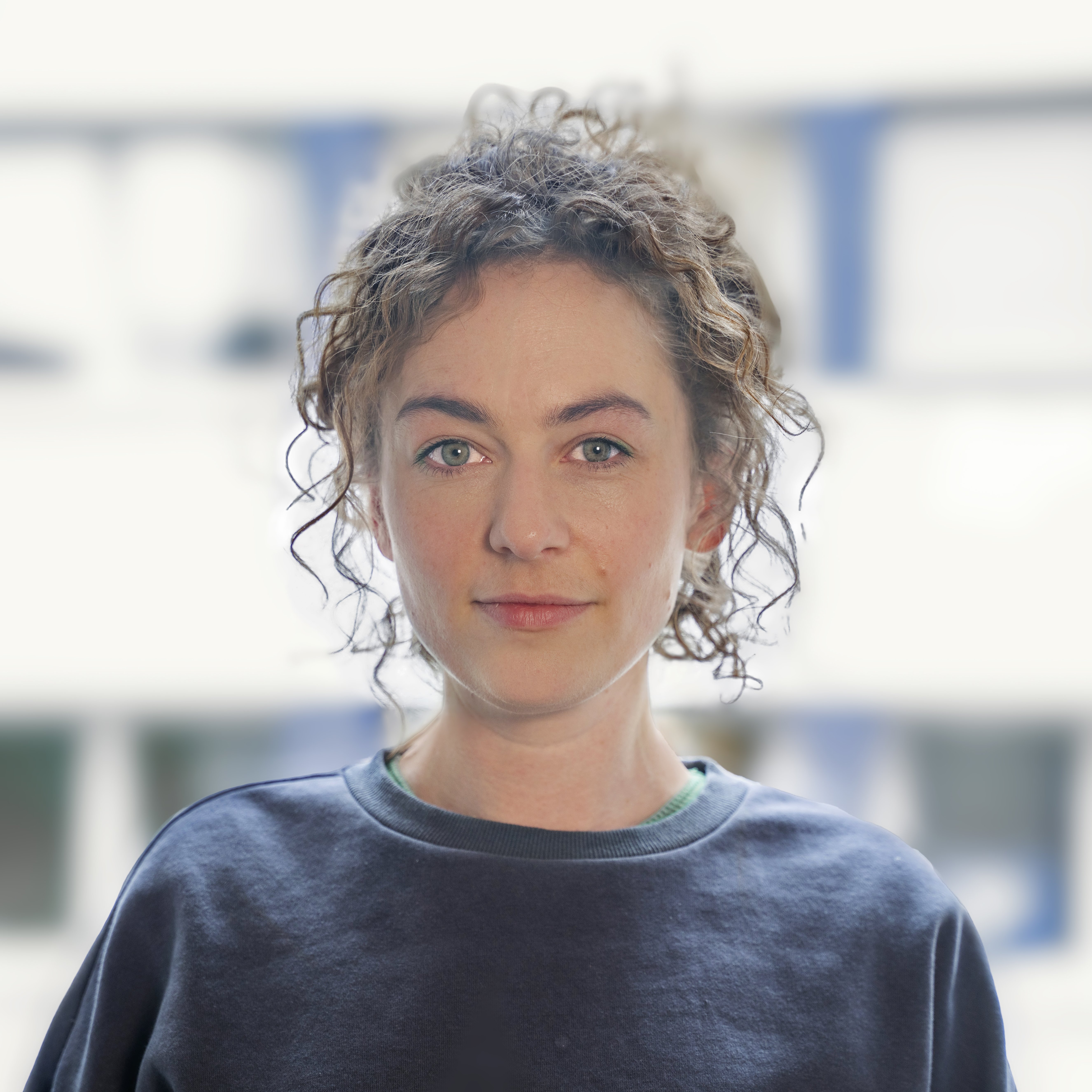 Portrait einer Person mit lockigem, braunem Haar, das zu einem hohen Pferdeschwanz gebunden ist. Sie hat grüne Augen und trägt einen dunkelblauen Pullover über einem grünen Hemd. Ihr Blick ist direkt auf den Betrachter gerichtet. Der Hintergrund ist unscharf und zeigt helle Flächen, die an Fenster oder Gebäude erinnern.