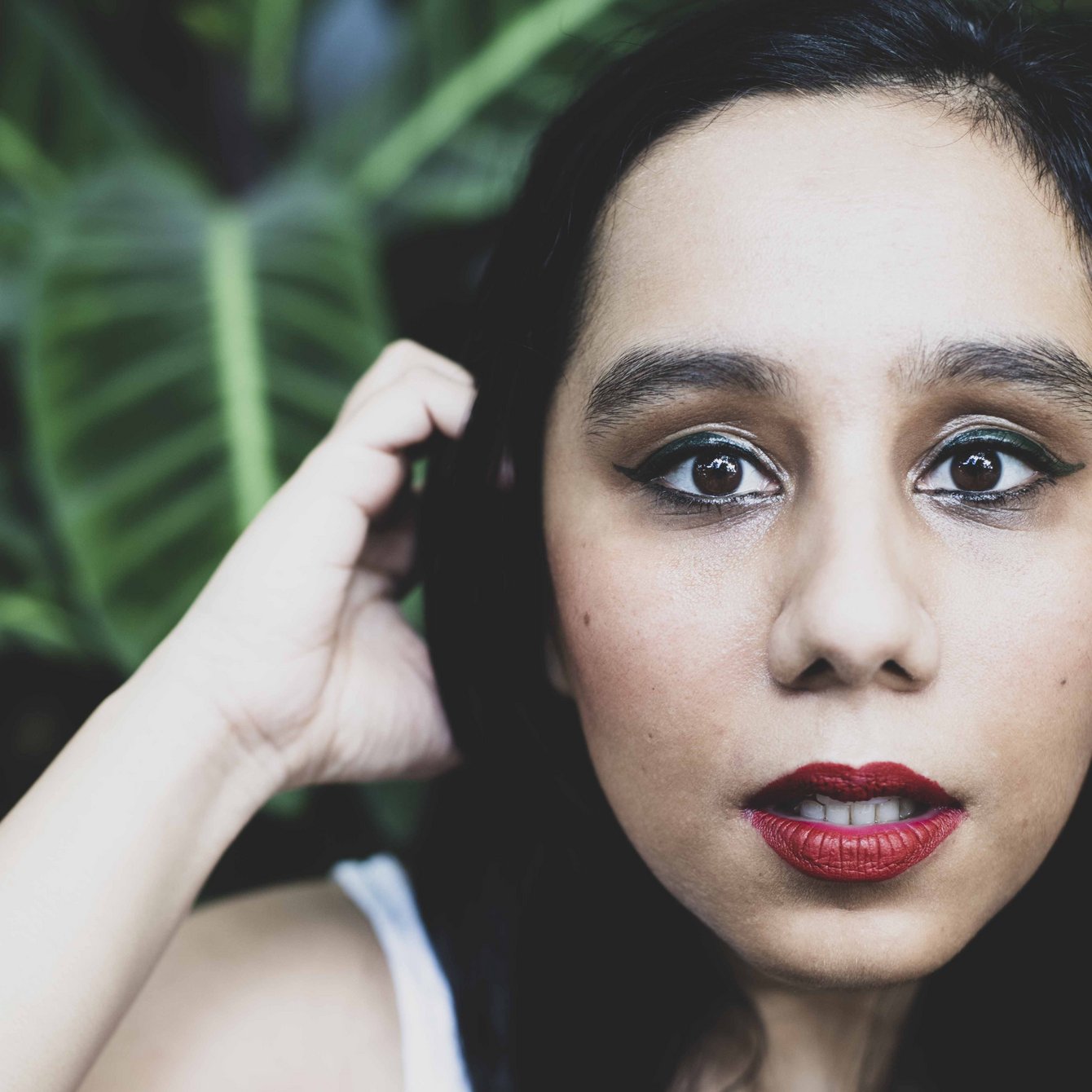 Portraitaufnahme von Deepika Arwind. Sie ist mit einem weißen ärmellosen Shirt bekleidet, trägt ihre langen schwarzen Haare offen und schiebt sich gerade mit der rechten Hand Haare aus dem Gesicht. Im Hintergrund ist eine grüne Pflanze zu sehen.