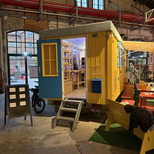 Ein gelber Bauwagen mit geöffneter Tür steht in einer Halle, umgeben von Tischen und Stühlen bei einem Kunst-Symposium.