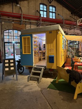 Ein gelber Bauwagen mit geöffneter Tür steht in einer Halle, umgeben von Tischen und Stühlen bei einem Kunst-Symposium.