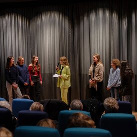 Moderatorin Viktoria Merkulova im Gespräch mit fünf Schüler*innen auf der Bühne vor der Kinoleinwand.