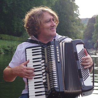 Peter Weiss hat lockige, mittellange Haare und spielt ein großes schwarzes Akkordeon der Marke Hohner, während er auf einem Boot auf einem Fluss sitzt. Er trägt ein helles Hemd und schaut lächelnd zur Seite. Im Hintergrund fahren weitere Menschen in einem Stocherkahn durch eine grüne, von Bäumen gesäumte Flusslandschaft.