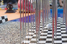 Minigolfball liegt auf einem schwarz-weiß karierten Spielfeld neben einem Hindernis aus senkrechten Metallstäben