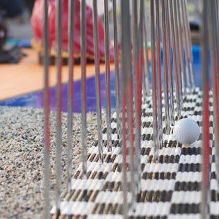 Minigolfball liegt auf einem schwarz-weiß karierten Spielfeld neben einem Hindernis aus senkrechten Metallstäben
