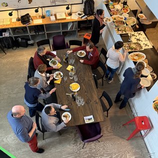 Die Gruppe beim Mittagessen: Mehrere Personen sitzen mit Tellern an einem Tisch. Rechts vom Tisch ist ein Buffet aufgebaut.